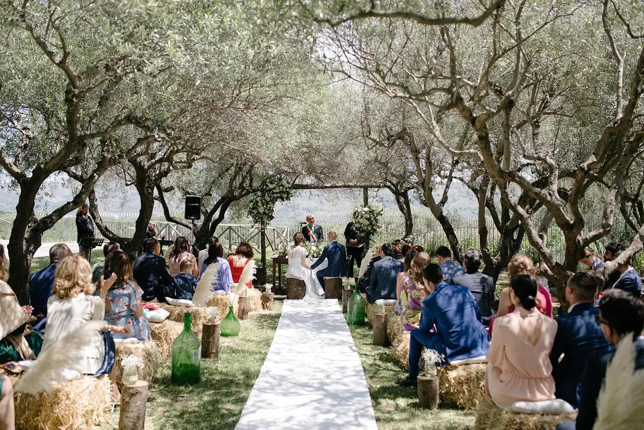 A ceremony under old olive trees at Borgata Baldazza, Linguaglossa, guests seated on hay bales along the aisle